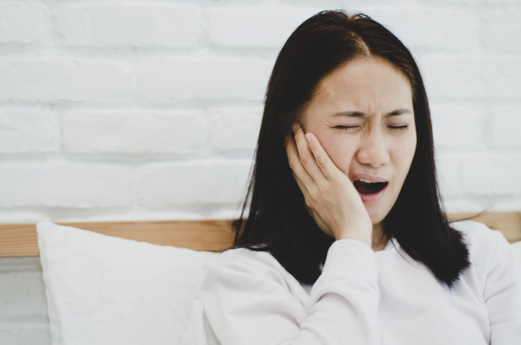 A sick woman sitting on a sofa is experiencing a toothache, her mouth is open while touching her jawline.