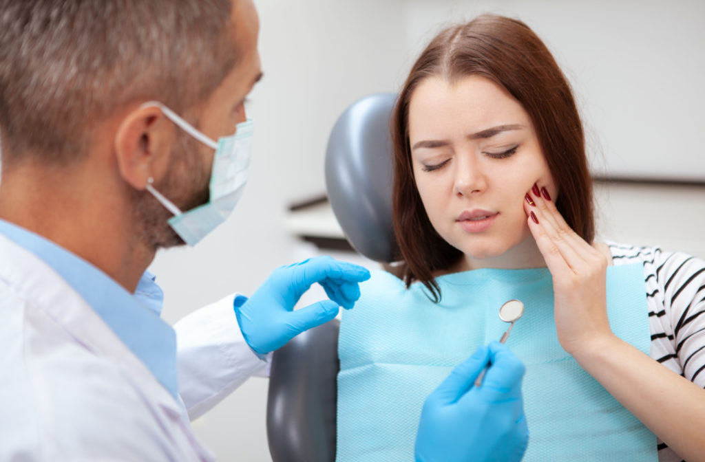 a woman sits in a dental chair holding her face with tooth pain. a dentist is preparing to examine her mouth
