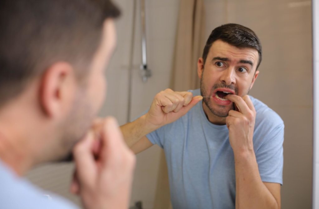 A man wincing in discomfort at the mirror while flossing his teeth.