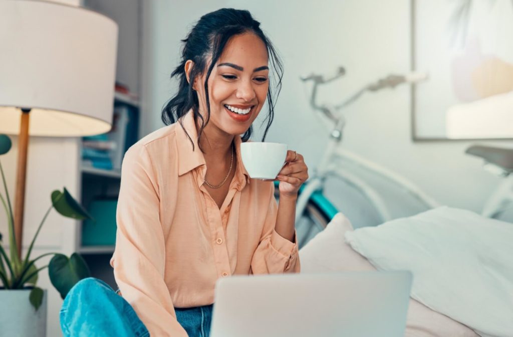 A smiling person working at their laptop is about to drink their cup of coffee.