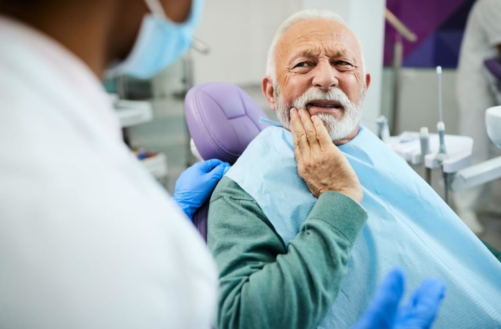 An older man in a dentist's office holds his jaw and describes his tooth pain.